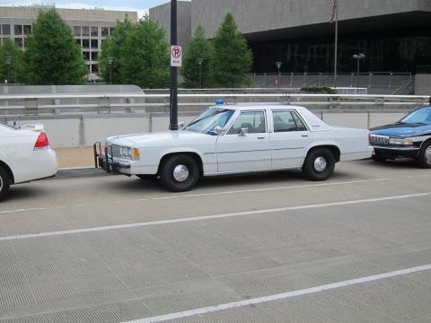 1991 Ford LTD Crown Victoria - white Ford LTD Crown Victoria police sedan with a push bar and blue beacon parked along a city street near a flag.