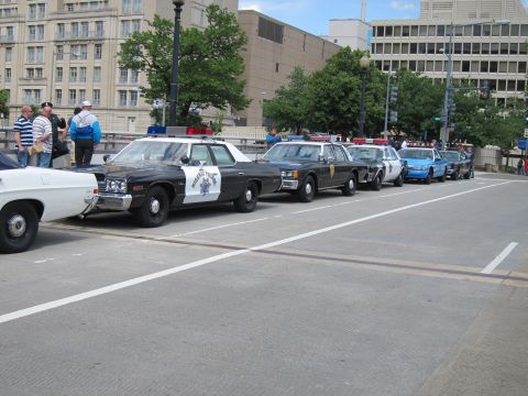 1991 Ford LTD Crown Victoria - lineup of classic black-and-white Highway Patrol and police cruisers parked along an urban street near tall buildings.