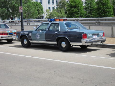 1991 Ford LTD Crown Victoria - two-tone blue State Police sedan with a lightbar and seal decal parked on a curbed urban street.
