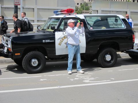 1991 Ford LTD Crown Victoria - man in a white shirt photographs a black-and-white Dodge Ram Charger SUV painted in Highway Patrol livery on a city street.