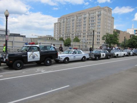 1991 Ford LTD Crown Victoria - row of marked police vehicles led by a black-and-white Highway Patrol SUV parked along a wide urban street.