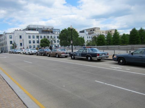 1991 Ford LTD Crown Victoria - line of older sedans and a dark blue marked police cruiser parked along a curb beneath cloudy skies.