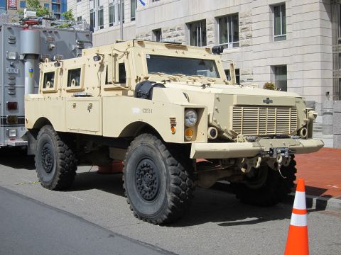 1991 Ford LTD Crown Victoria - tan armored MRAP-style four-wheel-drive vehicle with reinforced windows parked on an urban street near a traffic cone.