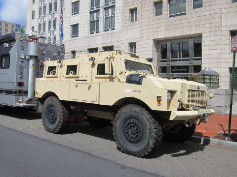 1991 Ford LTD Crown Victoria - tan armored MRAP-style vehicle with heavy off-road tires parked beside a silver command vehicle on a city street.
