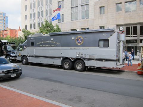 1991 Ford LTD Crown Victoria - gray FBI Mobile Command Center truck parked at a curb beneath U.S. flags on a downtown street.