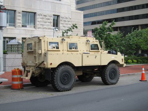 1991 Ford LTD Crown Victoria - tan armored MRAP-style vehicle with large off-road tires parked curbside near orange barrels and a traffic cone.