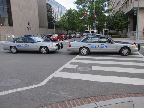 1991 Ford LTD Crown Victoria - Two silver State Police Ford sedans positioned at angles blocking a city intersection near a crosswalk.