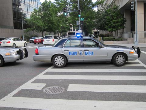 1991 Ford LTD Crown Victoria - silver State Police Ford sedan with blue lights crossing a striped crosswalk at a city intersection.
