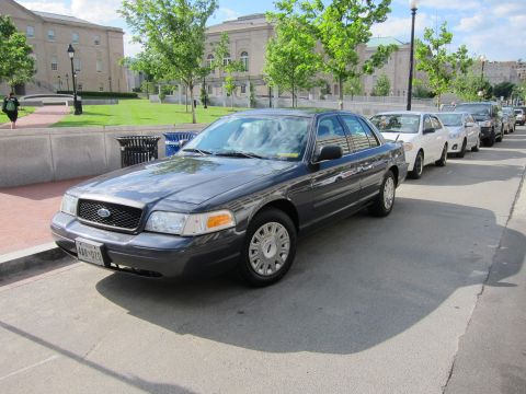 1991 Ford LTD Crown Victoria - dark gray Ford sedan with exempt plates parked at the front of a line of similar cars by a plaza.