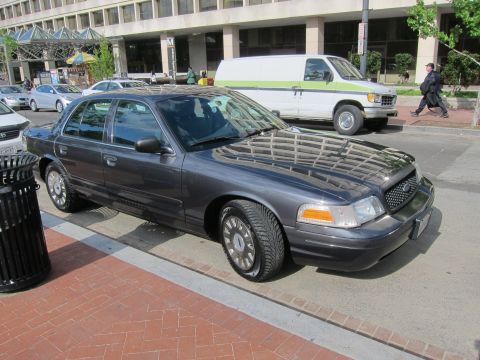 1991 Ford LTD Crown Victoria - gray Ford sedan parked at a brick sidewalk beside a trash can, with a green van in the background.
