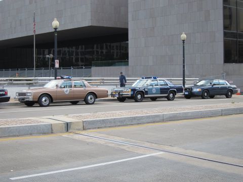 1991 Ford LTD Crown Victoria - brown vintage sedan, a blue and white police sedan, and a dark Ford sedan parked in a row by a curb.