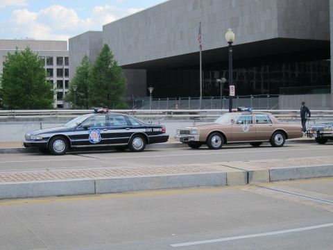 1991 Ford LTD Crown Victoria - black-and-white state trooper sedan parks beside a tan police cruiser on a city street in front of a large modernist concrete building.