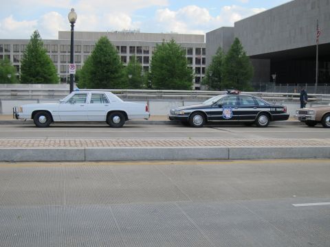 1991 Ford LTD Crown Victoria - white four-door sedan, a black police cruiser with door emblem, and a tan car line a curb beside a low office building.