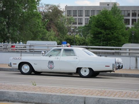 1991 Ford LTD Crown Victoria - white classic Dodge police sedan marked POLICE, with a blue dome light, sits curbside on a roadway with buildings behind.