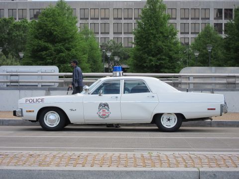 1991 Ford LTD Crown Victoria - man stands beside a white classic Dodge Fury police car marked POLICE, parked along a curb on a city street.