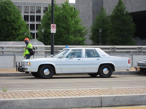 1991 Ford LTD Crown Victoria - white Ford LTD Crown Victoria police sedan with front push bar sits curbside as a worker in a hi-vis vest walks past.
