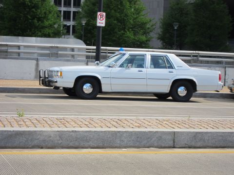 1991 Ford LTD Crown Victoria - white Ford LTD Crown Victoria police sedan with a chrome push bar and blue dome light parks beside a no-parking sign.