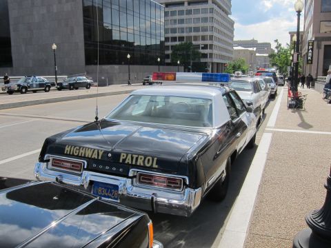 1991 Ford LTD Crown Victoria - rear of a black Dodge sedan lettered HIGHWAY PATROL leads a line of vintage police cars on a downtown street.