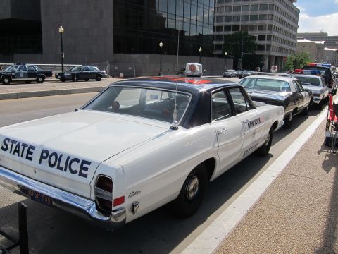 1991 Ford LTD Crown Victoria - white sedan lettered STATE POLICE with a NEW YORK door shield sits in a row of vintage cruisers on a downtown street.