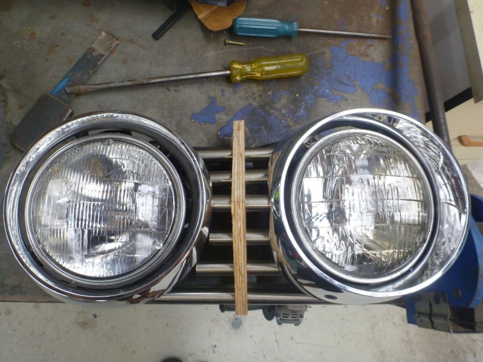 Chrome grille with two round headlights, held by a wooden plank. Screwdrivers and a metal tool are visible
