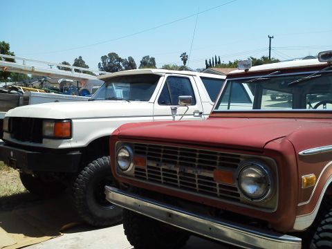 1988 Ford Bronco - Two SUVs, one red and one white, are parked next to each other. The red SUV's grille clearly displays the 'BRONCO' nameplate.