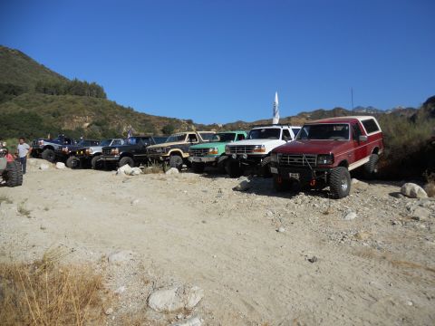 1986 Ford Bronco - A line of off-road vehicles, including a red and white Ford Bronco, are parked on a dirt lot under a clear blue sky.