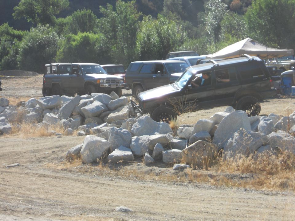 Dark SUV with roof rack driving over rocks, other SUVs and trucks in background
