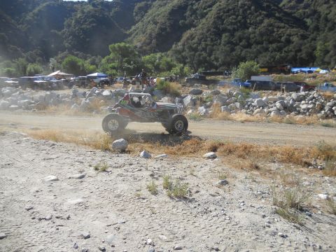 1986 Ford Bronco - An off-road buggy navigates a dusty track during an event, with spectators and other vehicles visible in the background.
