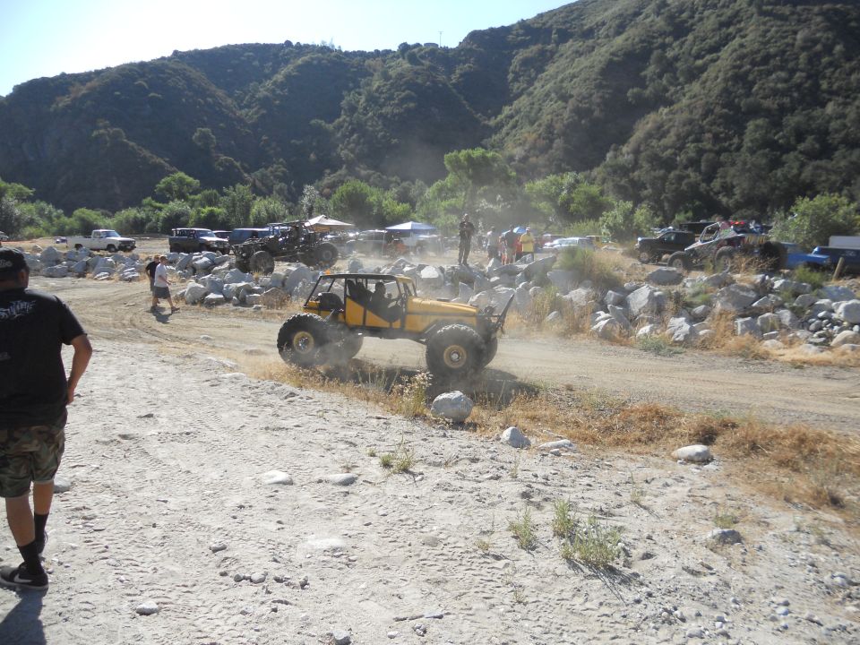 Yellow off-road buggy with large tires driving on a dirt path, kicking up dust. Mountains in background