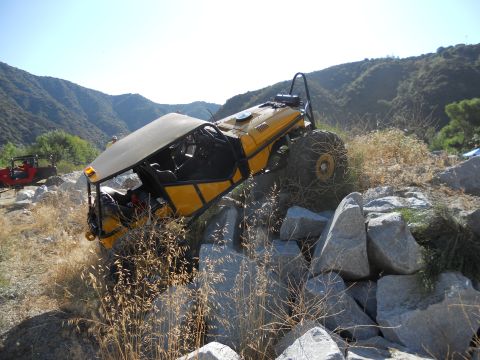 1986 Ford Bronco - A heavily modified yellow off-road buggy navigates a challenging rocky trail, with another red vehicle visible in the background.