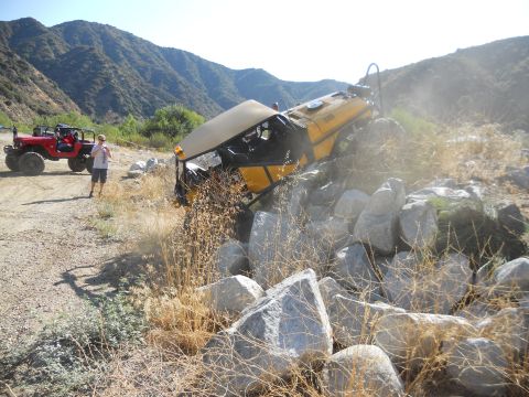 1986 Ford Bronco - An off-road vehicle is tipped on its side amidst rocks, with another Jeep and a person standing nearby in a mountainous landscape.