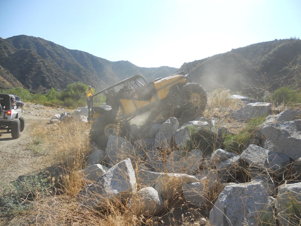 Yellow off-road buggy with large tires navigating a rocky incline, another white Jeep visible on the left