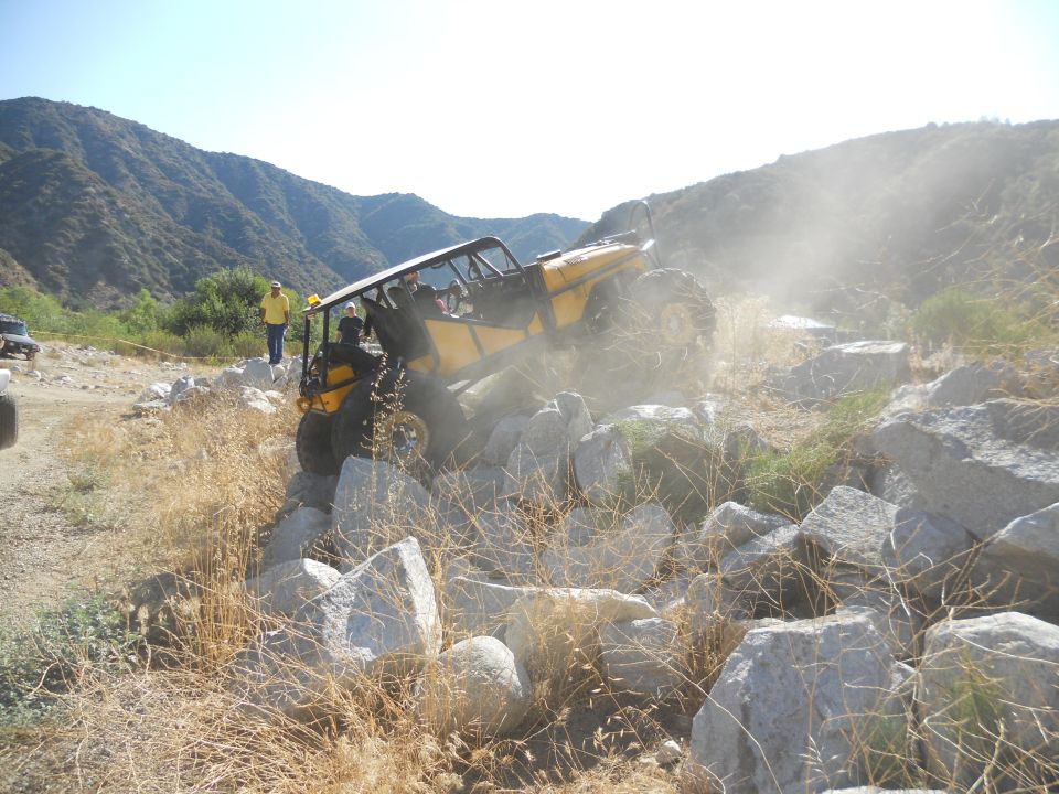 Yellow off-road buggy climbing over large rocks with dust in the air. Another vehicle is visible in the background