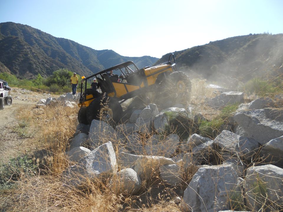 Yellow off-road buggy navigating a rocky trail, kicking up dust. Another white vehicle is visible in the background
