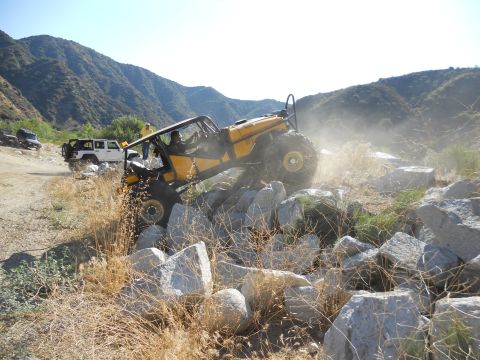 1986 Ford Bronco - An off-road vehicle navigates a rocky terrain, kicking up dust, with other vehicles visible in the distance.