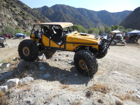1986 Ford Bronco - A heavily modified yellow off-road vehicle navigates a rocky trail with mountains in the background.