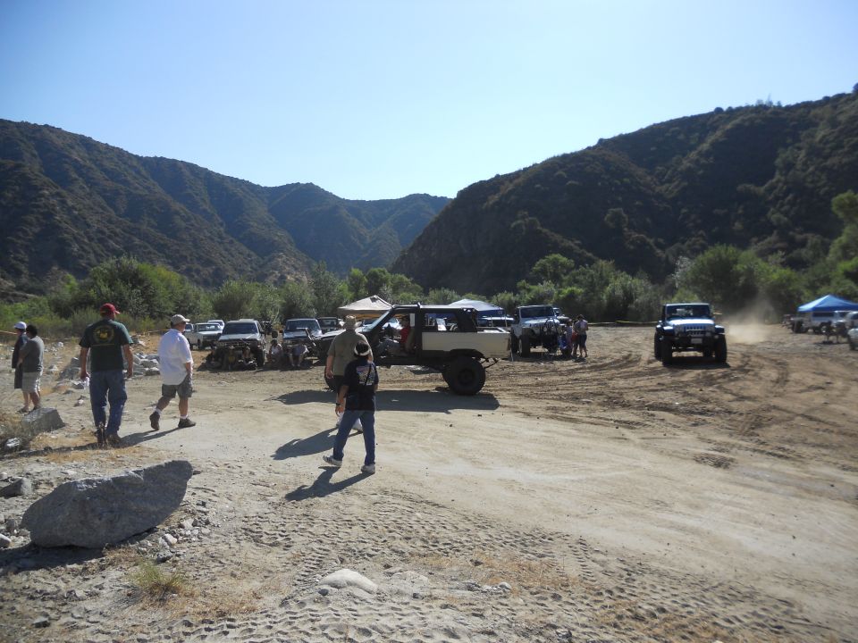 Several off-road vehicles parked and driving in a dusty, mountainous area. A group of people are gathered near some of the vehicles