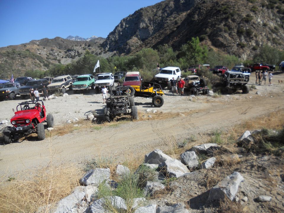 Red Jeep-like off-road vehicle in foreground, with many other off-road vehicles gathered in a dry, rocky, mountainous area