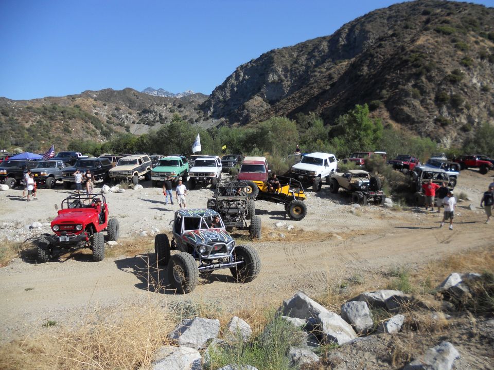 Many off-road vehicles parked in a dusty canyon with mountains in the background