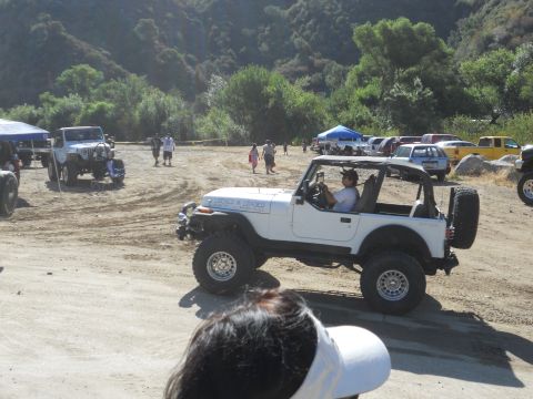 1986 Ford Bronco - A white Jeep Wrangler is seen in a sandy area with other vehicles and people in the background.