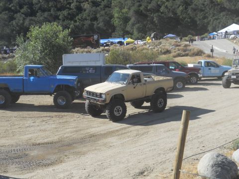 1986 Ford Bronco - A line of modified trucks are parked on a dusty lot, with a crowd and tents visible in the distance.