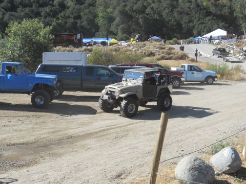 1986 Ford Bronco - Several off-road vehicles are parked in a dirt lot with tents and trees in the background.