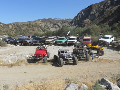 1986 Ford Bronco - A collection of modified off-road vehicles are gathered in a dusty, outdoor location with mountains in the background.