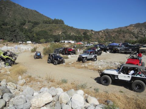 1986 Ford Bronco - A collection of off-road vehicles are parked and gathered on a dirt path, with a mountainous landscape visible behind them.