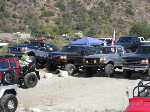 1986 Ford Bronco - A group of modified Ford trucks and SUVs are parked off-road, with one flying an American flag.