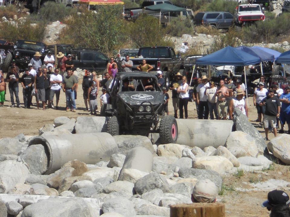 Off-road buggy on rocks, crowd watching. Other trucks and tents in background
