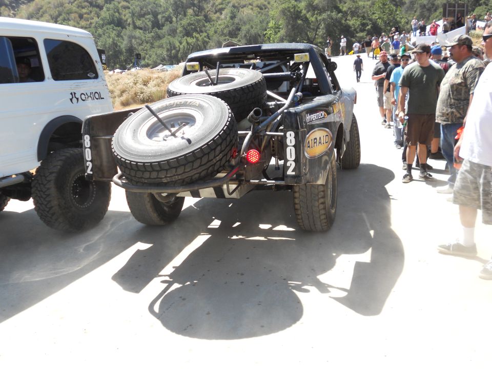 Off-road race truck with two spare tires mounted on the back, next to a white SUV