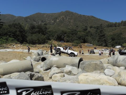 1986 Ford Bronco - Vehicles tackle a challenging off-road course with rocky terrain and large pipe obstacles under a clear blue sky.