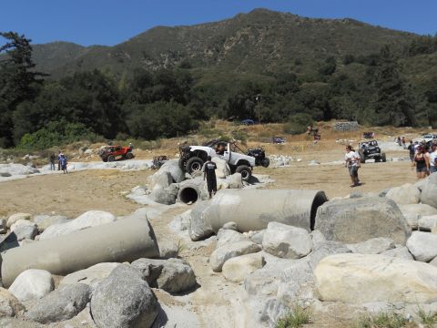 1986 Ford Bronco - Off-road vehicles tackle a challenging course with large rocks and concrete pipes under a clear blue sky.