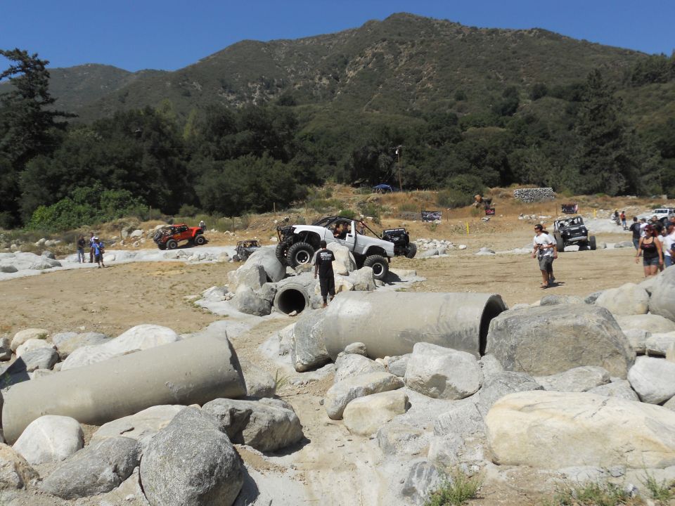 Several off-road vehicles, including a white Jeep-like vehicle and an orange buggy, are on a dirt course with large rocks and concrete pipes